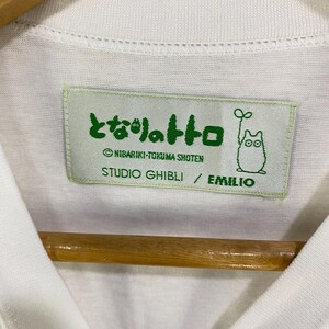 May include: White t-shirt with a Studio Ghibli label. The label is light green with Japanese text and a cartoon character illustration. The text includes "NIBARIKI-TOKUMA SHOTEN" and "STUDIO GHIBLI / EMILIO".