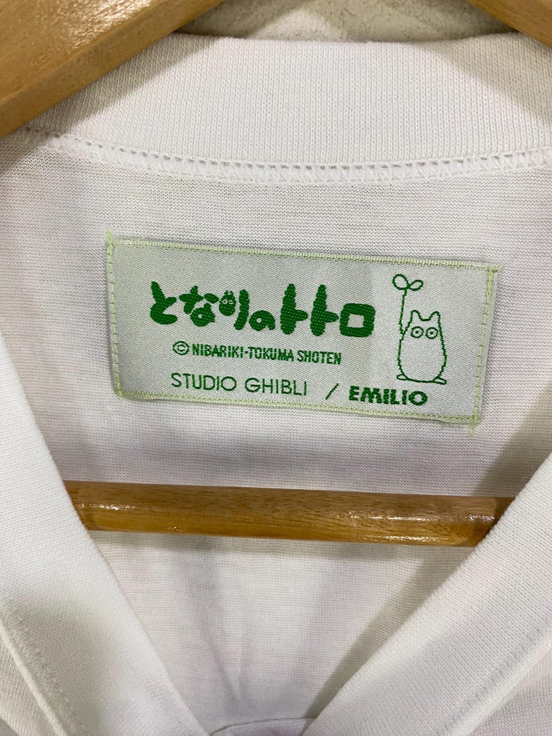 May include: White t-shirt with a Studio Ghibli label. The label is light green with Japanese text and a cartoon character illustration. The text includes "NIBARIKI-TOKUMA SHOTEN" and "STUDIO GHIBLI / EMILIO".