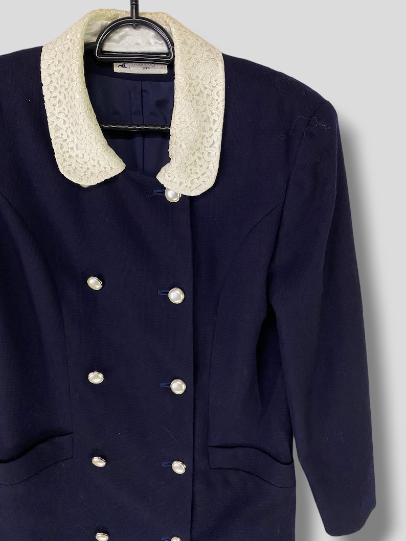 Puede incluir: Una chaqueta azul marino con cuello de encaje blanco y botones plateados. La chaqueta tiene cierre de doble botonadura y dos bolsillos.