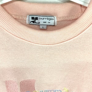 May include: A light pink Courr&egrave;ges and co sweatshirt. The label is white with black text and a logo. The sweatshirt has a ribbed crew neck and the size is L. The bottom of the sweatshirt has embroidered text in pastel colors.
