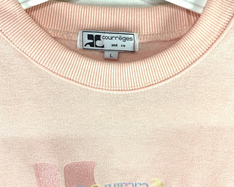 May include: A light pink Courr&egrave;ges and co sweatshirt. The label is white with black text and a logo. The sweatshirt has a ribbed crew neck and the size is L. The bottom of the sweatshirt has embroidered text in pastel colors.