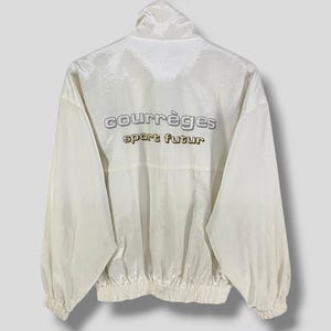 K&ouml;nnte beinhalten: Cremefarbene Jacke mit hohem Kragen und elastischen B&uuml;ndchen. Auf der R&uuml;ckseite der Jacke steht in silberner und goldener Schrift "courr&egrave;ges sport futur". Die Jacke h&auml;ngt an einem Metallb&uuml;gel.