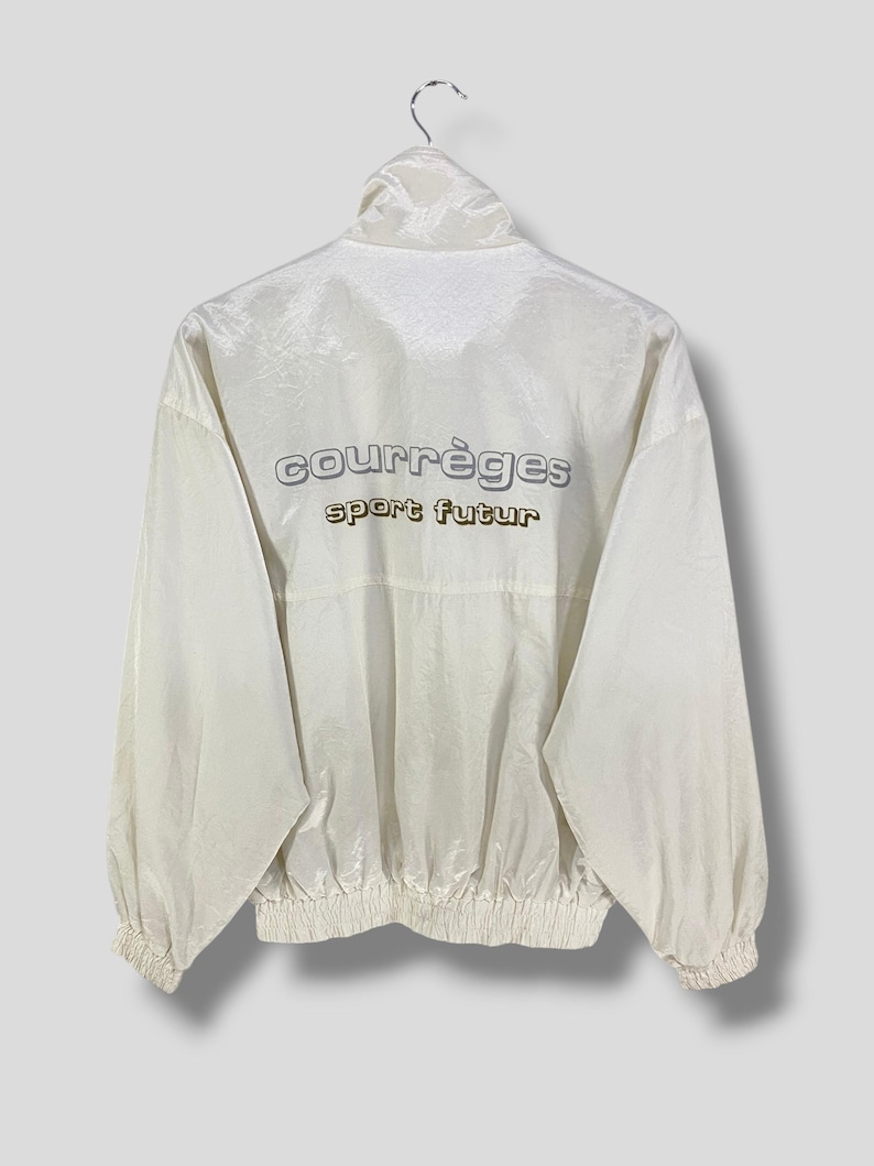 K&ouml;nnte beinhalten: Cremefarbene Jacke mit hohem Kragen und elastischen B&uuml;ndchen. Auf der R&uuml;ckseite der Jacke steht in silberner und goldener Schrift "courr&egrave;ges sport futur". Die Jacke h&auml;ngt an einem Metallb&uuml;gel.