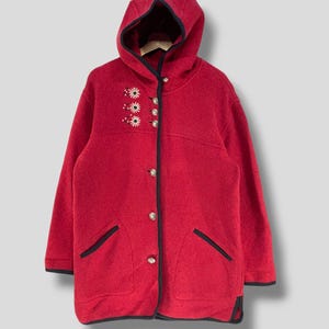 Può includere: Un cappotto rosso con cappuccio, finiture nere e dettagli floreali ricamati. Il cappotto presenta una chiusura con bottoni, due tasche anteriori e un cappuccio accogliente. La lunghezza del cappotto è di circa 76 cm.