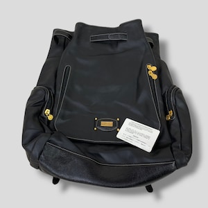 May include: Black leather backpack with gold-toned hardware, white stitching, and a drawstring closure. Features two side pockets with zippers and a small rectangular gold-toned emblem. A white tag with Japanese text is attached.