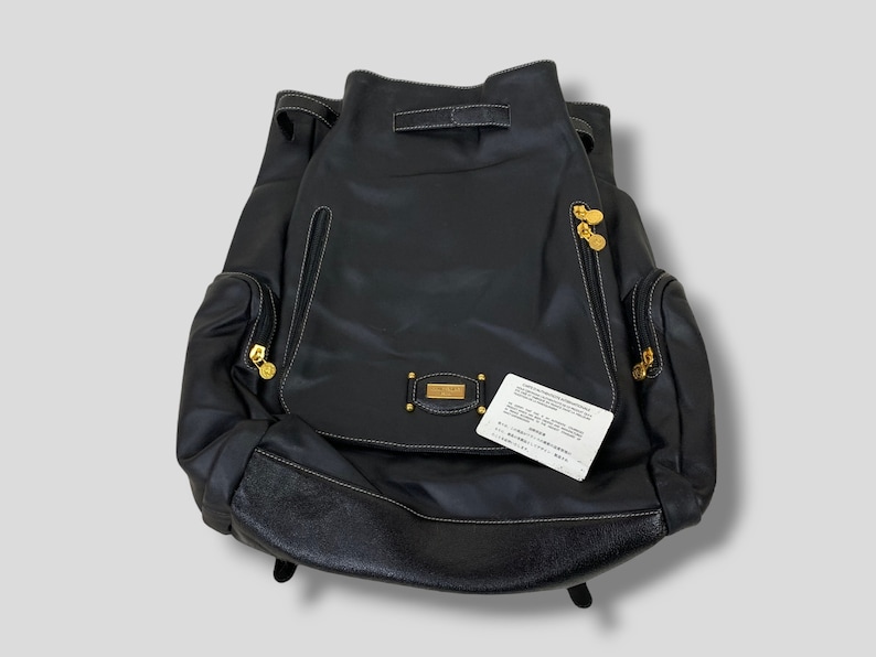 May include: Black leather backpack with gold-toned hardware, white stitching, and a drawstring closure. Features two side pockets with zippers and a small rectangular gold-toned emblem. A white tag with Japanese text is attached.