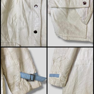 Puede incluir: Una chaqueta de cuero blanca con un detalle de correa azul en las mangas. La chaqueta tiene una cremallera y un bolsillo de solapa en la parte delantera.