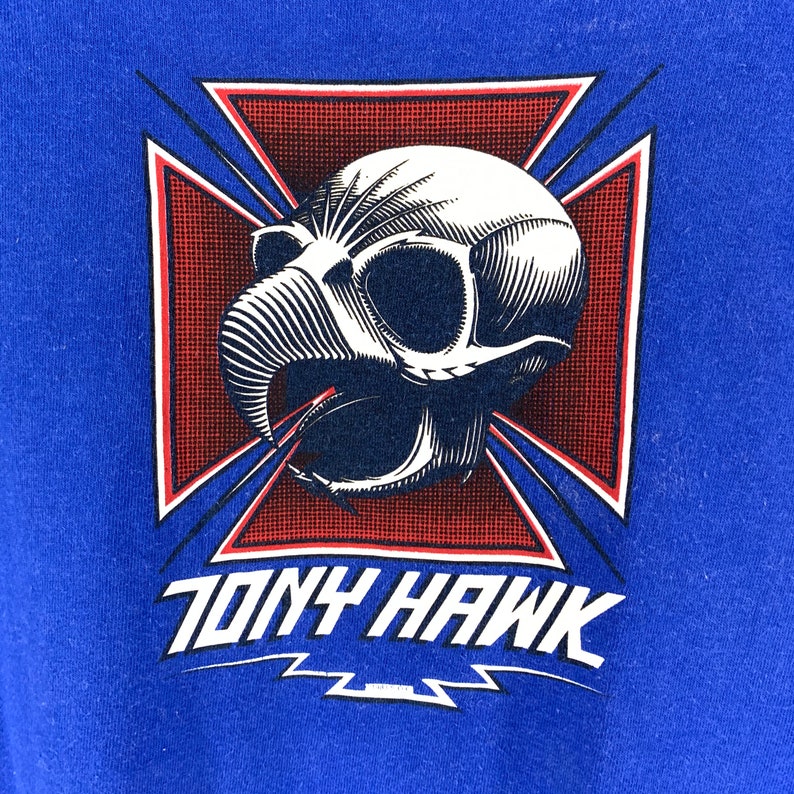 May include: Blue t-shirt with a graphic of a skull with a bird-like beak, set against a red and white cross design. The text "TONY HAWK" is printed in white below the graphic, with a lightning bolt design.
