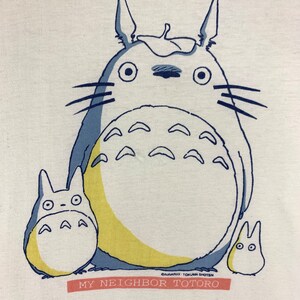 K&ouml;nnte beinhalten: Wei&szlig;es T-Shirt mit einer Grafik von Totoro und zwei kleineren Charakteren in Blau, Gelb und Wei&szlig;. Der Text "MY NEIGHBOR TOTORO" ist rot auf einem rechteckigen Banner darunter gedruckt.