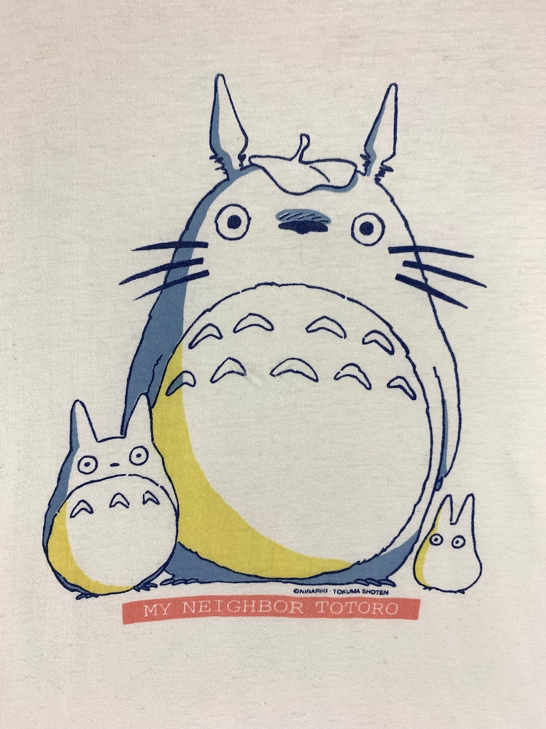 K&ouml;nnte beinhalten: Wei&szlig;es T-Shirt mit einer Grafik von Totoro und zwei kleineren Charakteren in Blau, Gelb und Wei&szlig;. Der Text "MY NEIGHBOR TOTORO" ist rot auf einem rechteckigen Banner darunter gedruckt.
