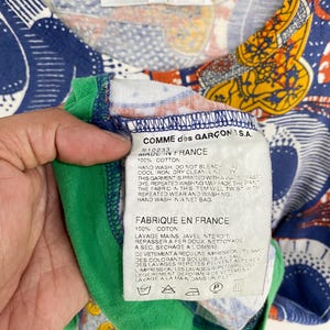 K&ouml;nnte beinhalten: Ein farbenfrohes Comme des Gar&ccedil;ons Shirt mit einem lebendigen Muster in Blau, Orange und Gelb. Das Shirt hat einen gr&uuml;nen Kragen und ein Etikett mit der Aufschrift "MADE IN FRANCE" und "100% COTTON". Gr&ouml;&szlig;e S.
