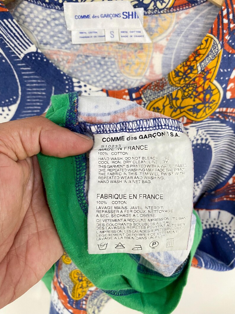 K&ouml;nnte beinhalten: Ein farbenfrohes Comme des Gar&ccedil;ons Shirt mit einem lebendigen Muster in Blau, Orange und Gelb. Das Shirt hat einen gr&uuml;nen Kragen und ein Etikett mit der Aufschrift "MADE IN FRANCE" und "100% COTTON". Gr&ouml;&szlig;e S.