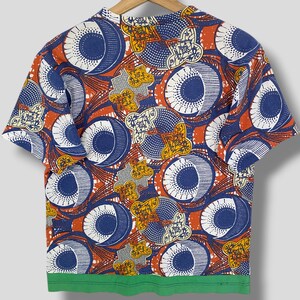 K&ouml;nnte beinhalten: Kurz&auml;rmeliges T-Shirt mit einem auff&auml;lligen Muster. Das Design zeigt gro&szlig;e blaue und wei&szlig;e kreisf&ouml;rmige Formen, orangefarbene und gelbe Akzente sowie einen gr&uuml;nen Saum. Das T-Shirt h&auml;ngt an einem Holzb&uuml;gel.