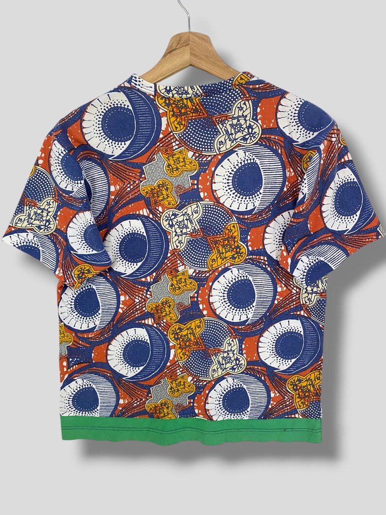 K&ouml;nnte beinhalten: Kurz&auml;rmeliges T-Shirt mit einem auff&auml;lligen Muster. Das Design zeigt gro&szlig;e blaue und wei&szlig;e kreisf&ouml;rmige Formen, orangefarbene und gelbe Akzente sowie einen gr&uuml;nen Saum. Das T-Shirt h&auml;ngt an einem Holzb&uuml;gel.