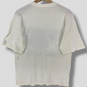 Puede incluir: Camiseta de color crema, de manga corta, colgada de una percha de madera. La camiseta tiene un dise&ntilde;o sencillo y cl&aacute;sico con cuello redondo y corte holgado. La tela parece suave y ligeramente desgastada, lo que sugiere un art&iacute;culo vintage o de segunda mano.