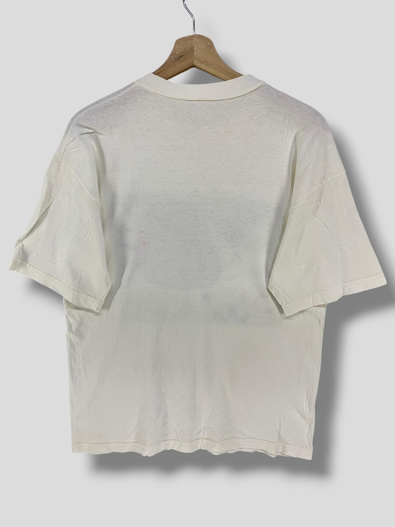 Puede incluir: Camiseta de color crema, de manga corta, colgada de una percha de madera. La camiseta tiene un dise&ntilde;o sencillo y cl&aacute;sico con cuello redondo y corte holgado. La tela parece suave y ligeramente desgastada, lo que sugiere un art&iacute;culo vintage o de segunda mano.