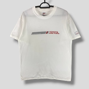 Könnte beinhalten: Weißes Kurzarm-T-Shirt mit dem Text "TOYOTA TEAM TOM'S" in Rot und einem grauen Streifen. Das Shirt hat auch den Text "THE VISION FOR MILLENNIUM" und ein kleines Toyota-Logo auf dem Ärmel.