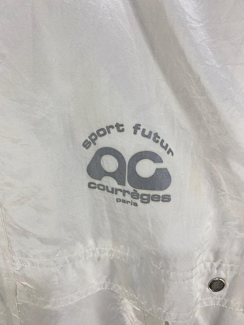 K&ouml;nnte beinhalten: Wei&szlig;e Nylonjacke mit dem Text "sport futur" und "courr&egrave;ges paris" in Grau. Die Jacke hat ein Logo mit den Buchstaben "ac" in einer stilisierten Schriftart. Sie hat einen Knopfverschluss.