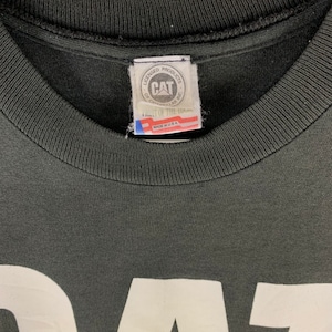 May include: Close-up of a black t-shirt with a white graphic. The shirt's collar and tag are visible. The tag reads "CAT" and "Made in USA". The shirt is made of a soft, comfortable material.