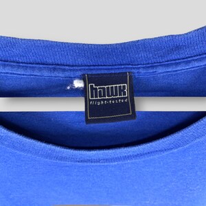 May include: Close-up of a blue t-shirt with a tag that reads "hawk flight-tested" in white lettering on a navy blue background. The shirt is made of a soft, comfortable material.