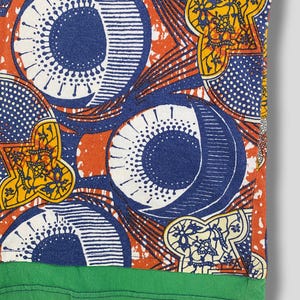 K&ouml;nnte beinhalten: Nahaufnahme eines gemusterten Stoffes mit gr&uuml;nem Saum. Das Design zeigt gro&szlig;e blaue Kreise, wei&szlig;e Punkte und gelbe Blumenformen auf orangefarbenem Hintergrund. Der Stoff scheint ein Abschnitt eines Hemdes oder eines anderen Kleidungsst&uuml;cks zu sein.