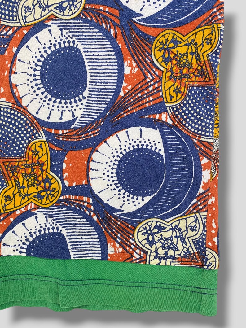 K&ouml;nnte beinhalten: Nahaufnahme eines gemusterten Stoffes mit gr&uuml;nem Saum. Das Design zeigt gro&szlig;e blaue Kreise, wei&szlig;e Punkte und gelbe Blumenformen auf orangefarbenem Hintergrund. Der Stoff scheint ein Abschnitt eines Hemdes oder eines anderen Kleidungsst&uuml;cks zu sein.