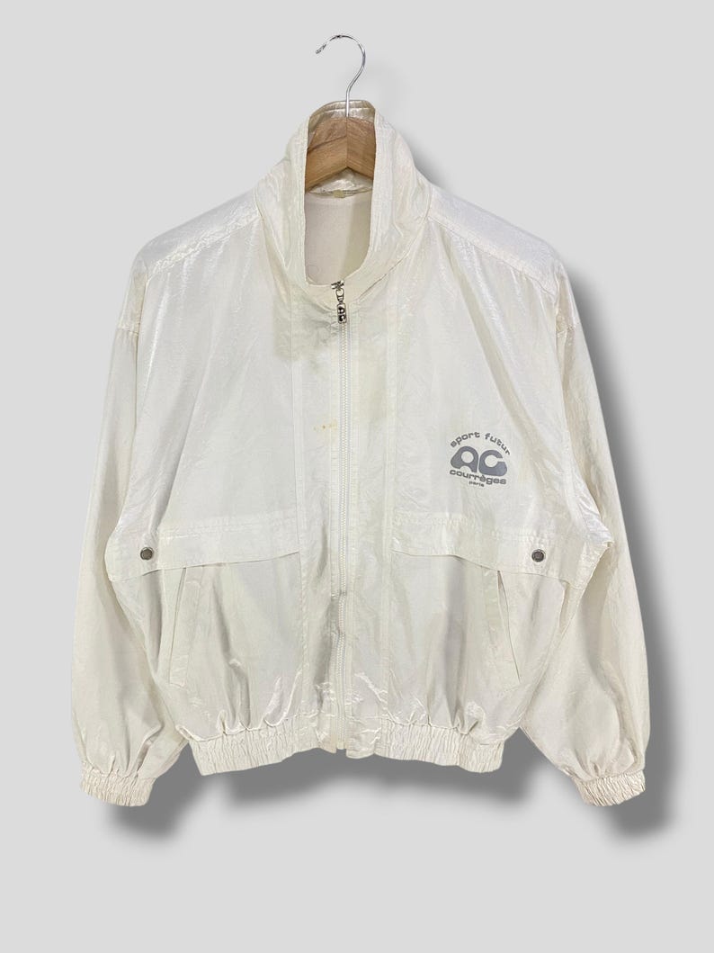 K&ouml;nnte beinhalten: Eine cremefarbene Vintage-Jacke mit hohem Kragen und durchgehendem Rei&szlig;verschluss. Die Jacke hat zwei Vordertaschen und ein Logo auf der rechten Brust mit der Aufschrift "sport futur" und "ac coutures". Die Jacke h&auml;ngt an einem Holzkleiderb&uuml;gel.