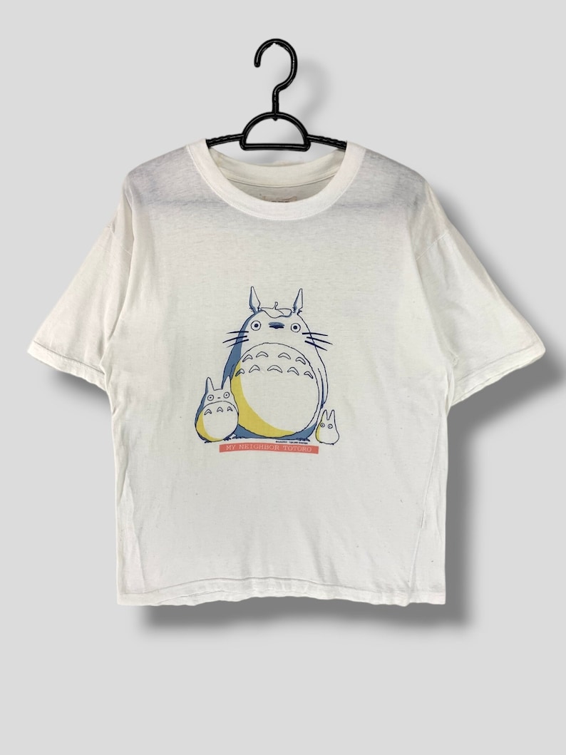 May include: White t-shirt with a cartoon image of Totoro from the movie My Neighbor Totoro. The image is in blue, yellow, and black. The text 'My Neighbor Totoro' is printed in red below the image.