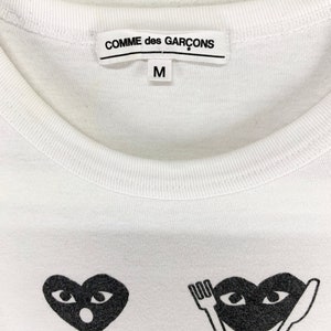 Puede incluir: Camiseta blanca de cuello redondo con el nombre de la marca "COMME des GAR&Ccedil;ONS" impreso en la etiqueta. La camiseta presenta dos gr&aacute;ficos de coraz&oacute;n negro con ojos. La talla "M" est&aacute; impresa en la etiqueta.