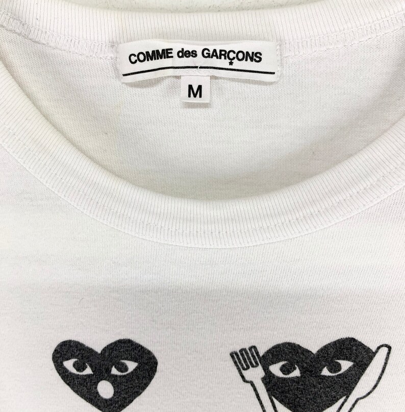 Puede incluir: Camiseta blanca de cuello redondo con el nombre de la marca "COMME des GAR&Ccedil;ONS" impreso en la etiqueta. La camiseta presenta dos gr&aacute;ficos de coraz&oacute;n negro con ojos. La talla "M" est&aacute; impresa en la etiqueta.