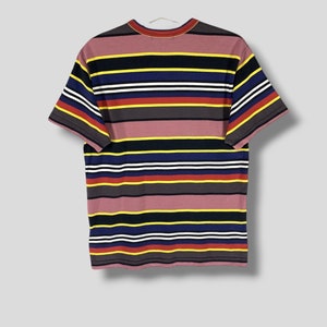 May include: A short-sleeved t-shirt with horizontal stripes in various colors, including mauve, yellow, navy, white, red, and gray. The shirt is hanging on a white hanger against a light gray background. The shirt's design is a classic, casual style.