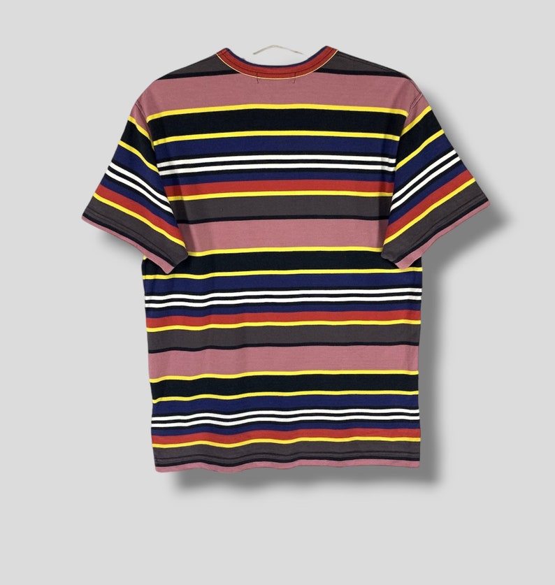 May include: A short-sleeved t-shirt with horizontal stripes in various colors, including mauve, yellow, navy, white, red, and gray. The shirt is hanging on a white hanger against a light gray background. The shirt's design is a classic, casual style.