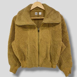 Può includere: Giacca sherpa color senape con chiusura a zip completa. La giacca presenta un ampio colletto, polsini e orlo a coste. La giacca è appesa a una gruccia di legno. Il materiale appare morbido e strutturato.