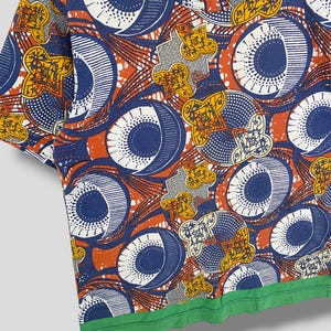K&ouml;nnte beinhalten: Ein kurz&auml;rmeliges T-Shirt mit Rundhalsausschnitt und einem auff&auml;lligen, sich wiederholenden Muster. Das Design umfasst gro&szlig;e blaue und wei&szlig;e Kreise, orangefarbene und gelbe Blumenformen sowie einen gr&uuml;nen Saum. Das T-Shirt besteht aus einem weichen, bequemen Material.