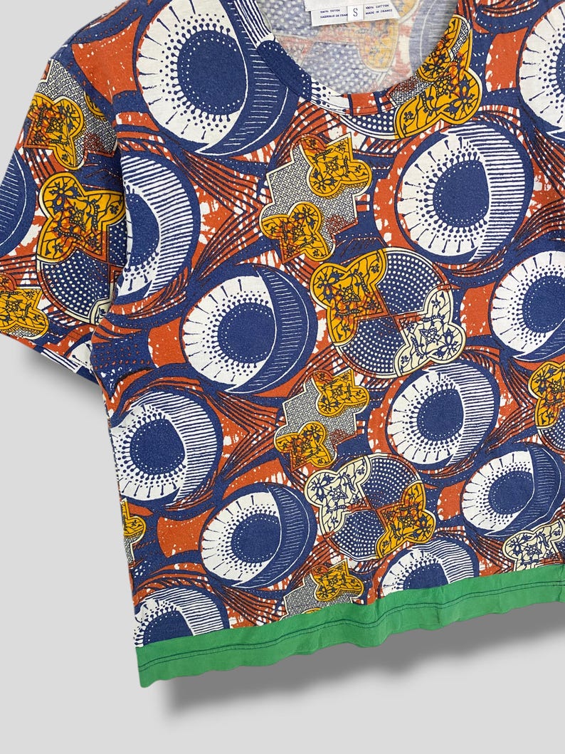 K&ouml;nnte beinhalten: Ein kurz&auml;rmeliges T-Shirt mit Rundhalsausschnitt und einem auff&auml;lligen, sich wiederholenden Muster. Das Design umfasst gro&szlig;e blaue und wei&szlig;e Kreise, orangefarbene und gelbe Blumenformen sowie einen gr&uuml;nen Saum. Das T-Shirt besteht aus einem weichen, bequemen Material.