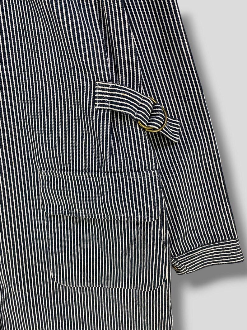 May include: A blue and white striped workwear jacket with a large pocket on the front and a metal ring on the sleeve.