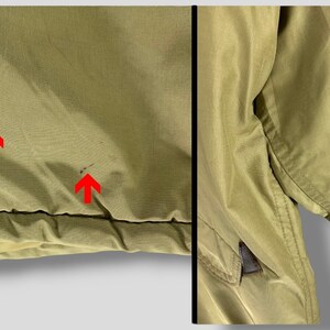 Puede incluir: Primer plano de una chaqueta verde oliva claro. La chaqueta muestra algunas manchas e imperfecciones menores. La tela parece ser un material ligero, posiblemente nailon o una mezcla sint&eacute;tica similar. La chaqueta tiene un dise&ntilde;o simple y utilitario.
