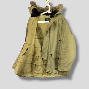 Puede incluir: Abrigo de invierno verde oliva con capucha forrada de piel, colgado de una percha blanca. El abrigo tiene un interior acolchado y m&uacute;ltiples bolsillos. El abrigo est&aacute; abotonado y tiene cierre de cremallera. El abrigo es de material resistente al agua.