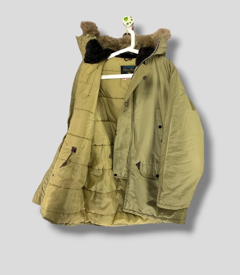 Puede incluir: Abrigo de invierno verde oliva con capucha forrada de piel, colgado de una percha blanca. El abrigo tiene un interior acolchado y m&uacute;ltiples bolsillos. El abrigo est&aacute; abotonado y tiene cierre de cremallera. El abrigo es de material resistente al agua.