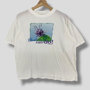 Puede incluir: Camiseta blanca con un personaje de dibujos animados, una hormiga de la película "Bichos". La hormiga es morada con ojos grandes y alas, de pie sobre una hoja verde. El texto "Blueberry can-do!" está debajo.
