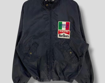 May include: A black nylon jacket with a stand-up collar and a full-length zipper. The jacket features two front pockets and patches with the text "J.ALESI" and "Marlboro" along with a flag design. The jacket is hanging on a wooden hanger.