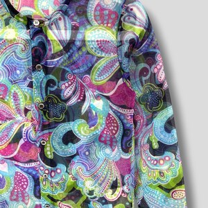 Puede incluir: Una camisa transparente con botones y un vibrante estampado de cachemira en tonos azules, morados, verdes y rosas. La camisa presenta un cuello mandar&iacute;n y varios botones peque&ntilde;os en la parte delantera. La etiqueta dice "tricot COMME des GAR&Ccedil;ONS."