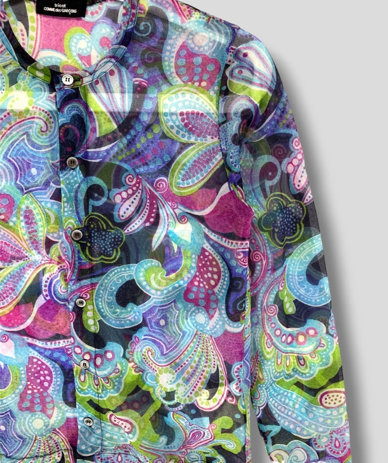 Puede incluir: Una camisa transparente con botones y un vibrante estampado de cachemira en tonos azules, morados, verdes y rosas. La camisa presenta un cuello mandar&iacute;n y varios botones peque&ntilde;os en la parte delantera. La etiqueta dice "tricot COMME des GAR&Ccedil;ONS."