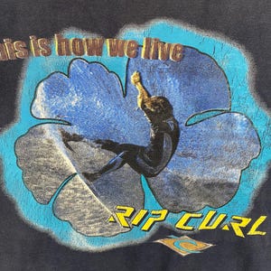May include: Dark blue t-shirt featuring a surfer inside a blue flower design. The text "this is how we live" is arched above the flower, and "RIP CURL" is below. The Rip Curl logo is at the bottom.