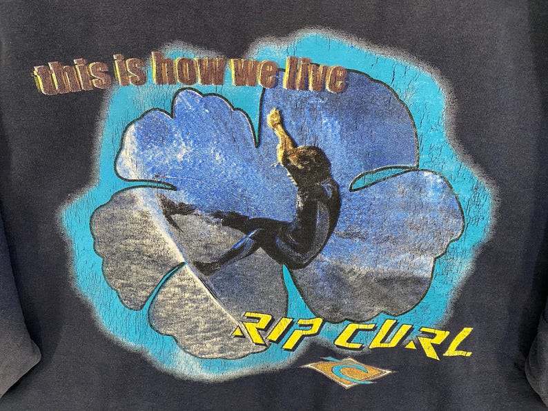 May include: Dark blue t-shirt featuring a surfer inside a blue flower design. The text "this is how we live" is arched above the flower, and "RIP CURL" is below. The Rip Curl logo is at the bottom.
