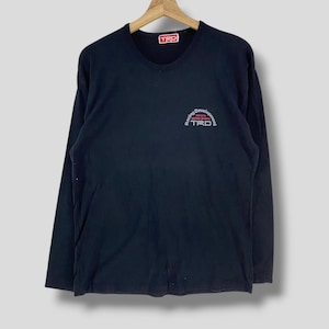 May include: A navy blue long-sleeve shirt with a crew neck. The shirt features the text "TRD" in a red rectangle at the top and a logo with the text "Racing Development" and "TRD" in red and white on the chest. The shirt is hanging on a wooden hanger.