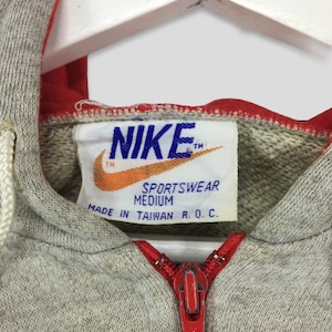 K&ouml;nnte beinhalten: Nahaufnahme eines grauen Nike Sportswear Hoodies mit rotem Rei&szlig;verschluss und Futter. Das Nike-Logo ist auf einem Etikett zu sehen, zusammen mit dem Text "Sportswear Medium Made in Taiwan R.O.C."