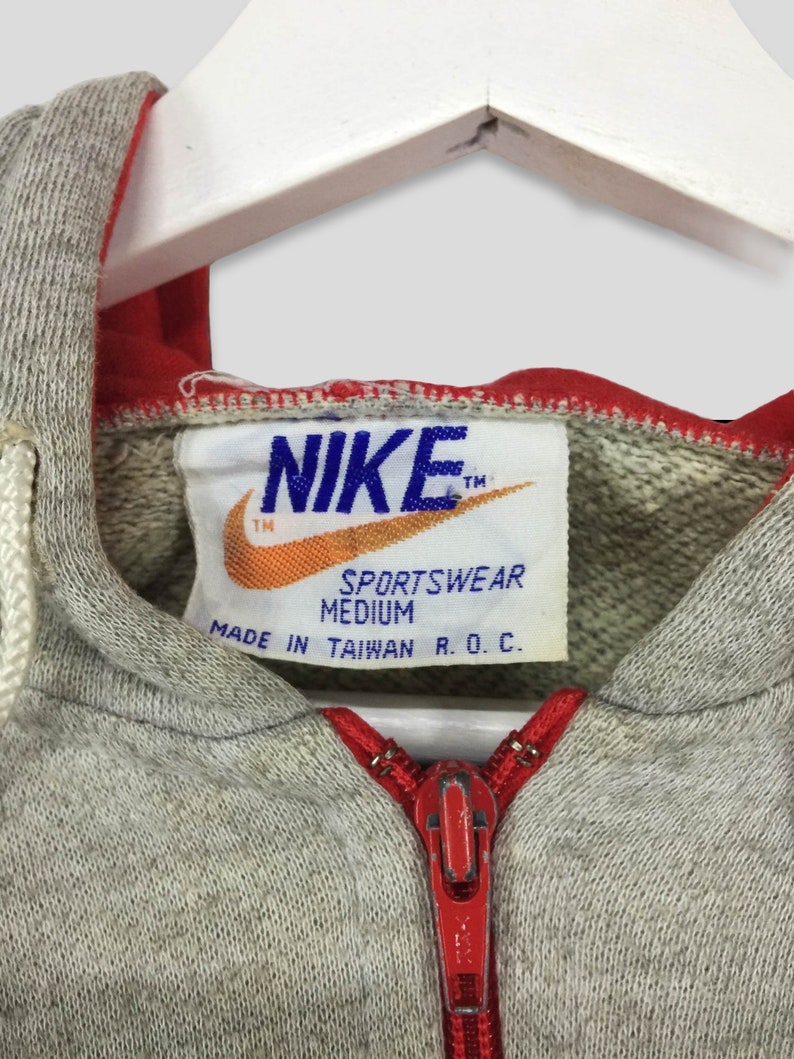 K&ouml;nnte beinhalten: Nahaufnahme eines grauen Nike Sportswear Hoodies mit rotem Rei&szlig;verschluss und Futter. Das Nike-Logo ist auf einem Etikett zu sehen, zusammen mit dem Text "Sportswear Medium Made in Taiwan R.O.C."