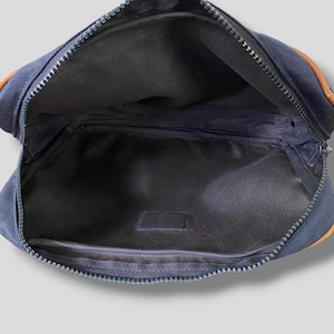 May include: Navy blue fanny pack with a zipper closure. The bag has brown leather accents on the corners. The interior is black.