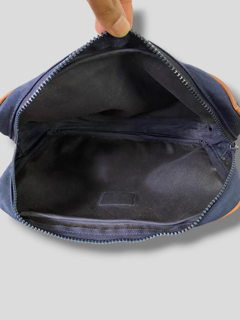 May include: Navy blue fanny pack with a zipper closure. The bag has brown leather accents on the corners. The interior is black.